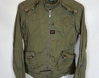 May include: Olive green jacket with multiple pockets, a zip-up front, and a small logo patch. The jacket has a stand-up collar and is hanging on a wooden hanger. The jacket is made of a durable material.