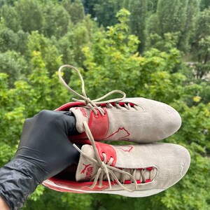 May include: Pair of Puma sneakers in a light gray suede material with red accents. The sneakers feature red Puma logos and laces. The shoes are held up against a backdrop of green trees.