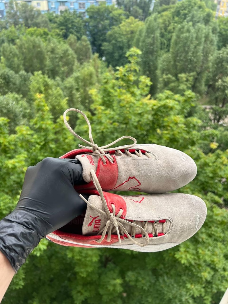 May include: Pair of Puma sneakers in a light gray suede material with red accents. The sneakers feature red Puma logos and laces. The shoes are held up against a backdrop of green trees.