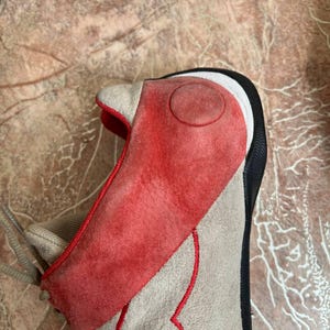 May include: A close-up of a sneaker with a unique design. The shoe features a red suede overlay on the toe and side, with a tan suede base and red stitching. The heel is black and white.