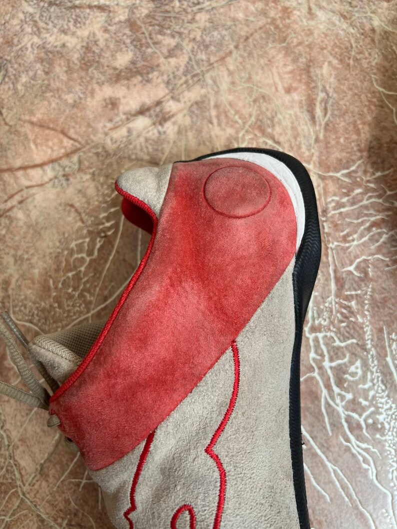 May include: A close-up of a sneaker with a unique design. The shoe features a red suede overlay on the toe and side, with a tan suede base and red stitching. The heel is black and white.