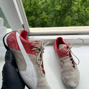 May include: Pair of Puma sneakers in gray, red, and white. The shoes have a suede-like texture, with a white Puma logo on the side. The red accents are on the top and heel. The sneakers are laced up.