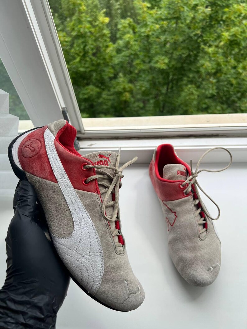 May include: Pair of Puma sneakers in gray, red, and white. The shoes have a suede-like texture, with a white Puma logo on the side. The red accents are on the top and heel. The sneakers are laced up.