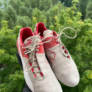 May include: A pair of Puma sneakers in a neutral beige suede with red accents. The shoes feature red detailing around the laces and heel, with the Puma logo visible. The soles are white, and the laces are beige.