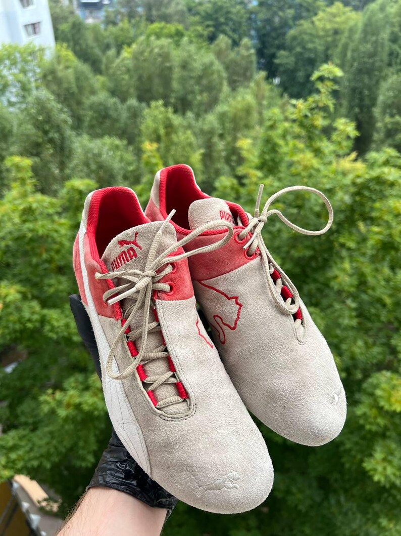 May include: A pair of Puma sneakers in a neutral beige suede with red accents. The shoes feature red detailing around the laces and heel, with the Puma logo visible. The soles are white, and the laces are beige.