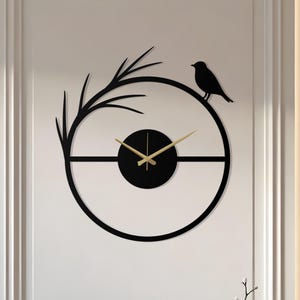 Op de afbeelding: Een zwarte metalen wandklok met een vogel- en takontwerp. De klok heeft een cirkelvorm met een zwart midden en goudkleurige wijzers. De vogel zit rechtsboven op de klok en de tak linksboven.