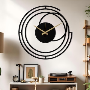 May include: A large, modern wall clock with a black circular face and gold-colored hands. The clock is set against a white wall and features a unique, geometric black frame. The design includes a black circle in the center and curved lines.