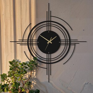 Puede incluir: Un reloj de pared negro y moderno con manecillas doradas y una esfera circular. El reloj presenta un diseño geométrico con líneas que se cruzan y círculos concéntricos. El reloj está montado en una pared texturizada de color claro, con una planta verde en primer plano.