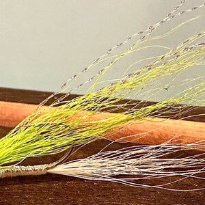 Op de afbeelding: Een groene en witte vliegvislure met een rode kraal. De lure is gebonden met verschillende materialen, waaronder veren en garen.