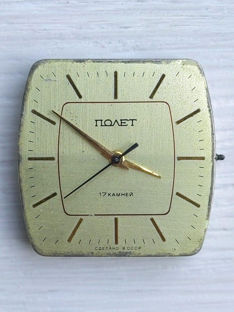 May include: A vintage square clock face with a gold-colored finish. The clock face features the word "ПОЛЕТ" in Cyrillic script, along with the text "17 KAMHEЙ" and "СДЕЛАНО В СССР". The clock hands are gold and black.