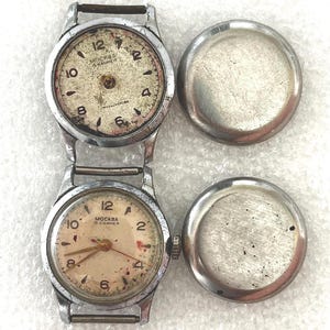 Puede incluir: Dos relojes de pulsera vintage plateados con esferas color crema y números negros. Las esferas muestran el texto "MOCKBA 16 KAMHEЙ". Los relojes se muestran sin sus traseras.