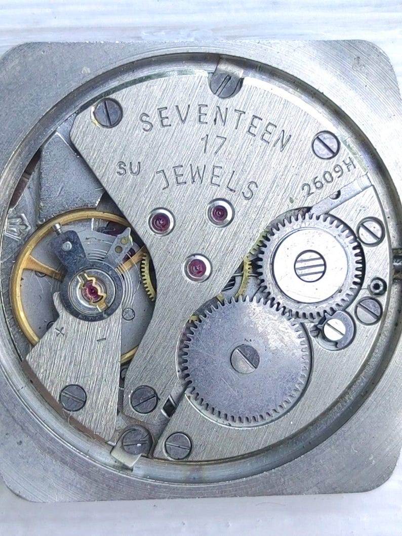 May include: Close-up of a watch movement with visible gears, screws, and the text "SEVENTEEN 17 JEWELS". The mechanism is housed in a square, silver-colored frame. The intricate design showcases the internal workings of a timepiece.