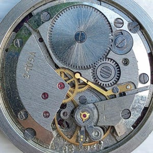 May include: Close-up of a watch movement with intricate gears, screws, and metal components. The silver-colored mechanism features a large central gear and smaller, interlocking gears. The text "2409A" is visible on a metal plate.