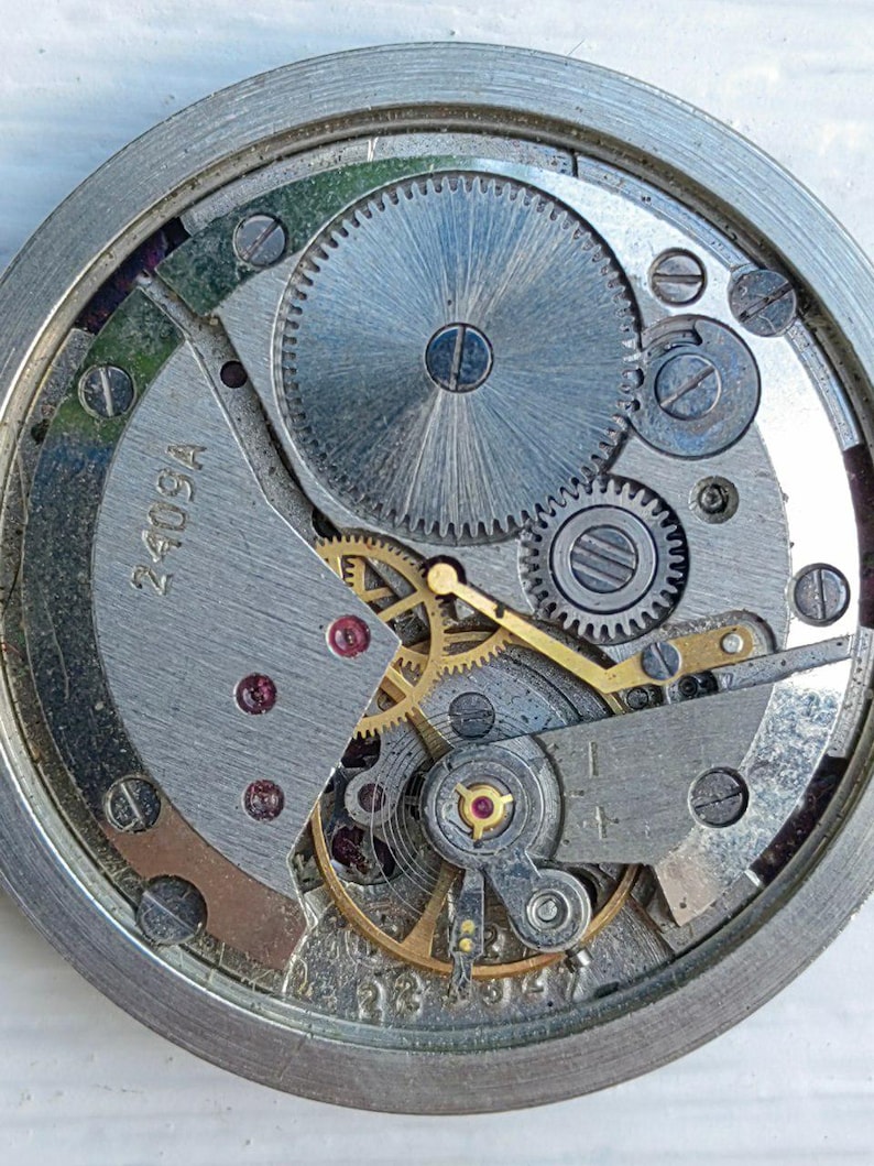 May include: Close-up of a watch movement with intricate gears, screws, and metal components. The silver-colored mechanism features a large central gear and smaller, interlocking gears. The text "2409A" is visible on a metal plate.