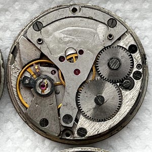 May include: Close-up of a vintage watch movement, showcasing intricate gears, springs, and balance wheel. The metal components are silver and gold, with red jewels. The circular mechanism is a detailed example of horological engineering.