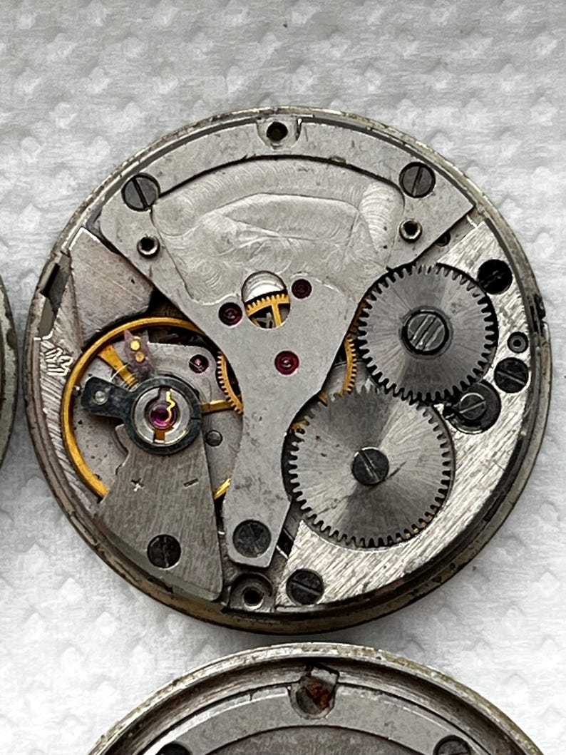 May include: Close-up of a vintage watch movement, showcasing intricate gears, springs, and balance wheel. The metal components are silver and gold, with red jewels. The circular mechanism is a detailed example of horological engineering.
