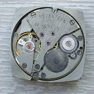 K&ouml;nnte beinhalten: Nahaufnahme eines Vintage-Uhrwerks mit sichtbaren Zahnr&auml;dern, Federn und Juwelen. Die Metallkomponenten sind silberfarben, mit dem Text "SEVENTEEN JEWELS" und "26098" sichtbar. Der Uhrenmechanismus ist quadratisch.