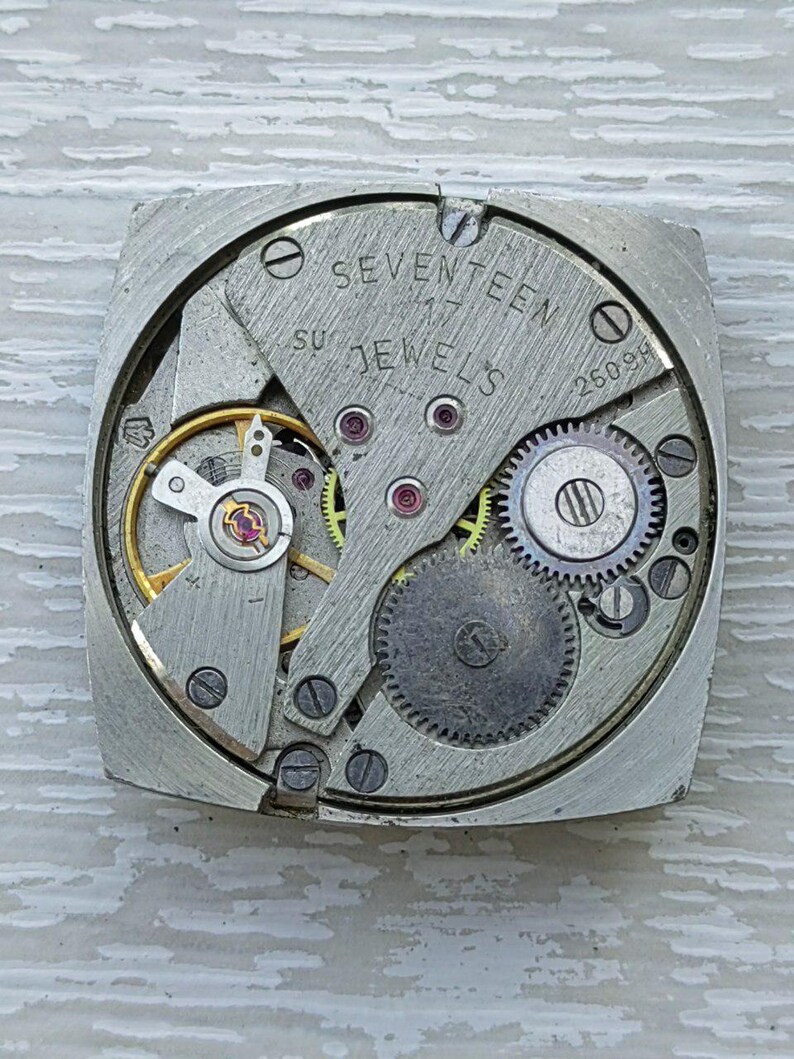 K&ouml;nnte beinhalten: Nahaufnahme eines Vintage-Uhrwerks mit sichtbaren Zahnr&auml;dern, Federn und Juwelen. Die Metallkomponenten sind silberfarben, mit dem Text "SEVENTEEN JEWELS" und "26098" sichtbar. Der Uhrenmechanismus ist quadratisch.
