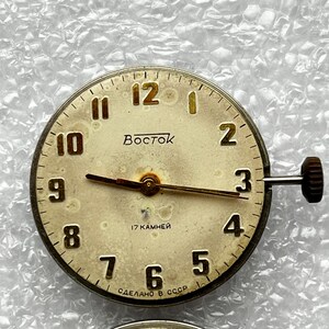 May include: A vintage watch face with a cream-colored dial, featuring black numerals and hands. The watch face has the word "Восток" and the text "17 KAMHEЙ" and "СДЕЛАНО В СССР". The watch has a crown on the side.