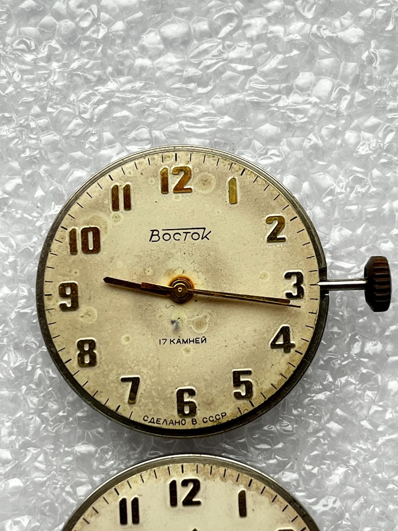 May include: A vintage watch face with a cream-colored dial, featuring black numerals and hands. The watch face has the word "Восток" and the text "17 KAMHEЙ" and "СДЕЛАНО В СССР". The watch has a crown on the side.