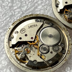 May include: Close-up of two disassembled watch movements. The watch mechanisms feature intricate gears, screws, and a balance wheel. The main plate is marked with "2409A". The components are silver and gold in color.