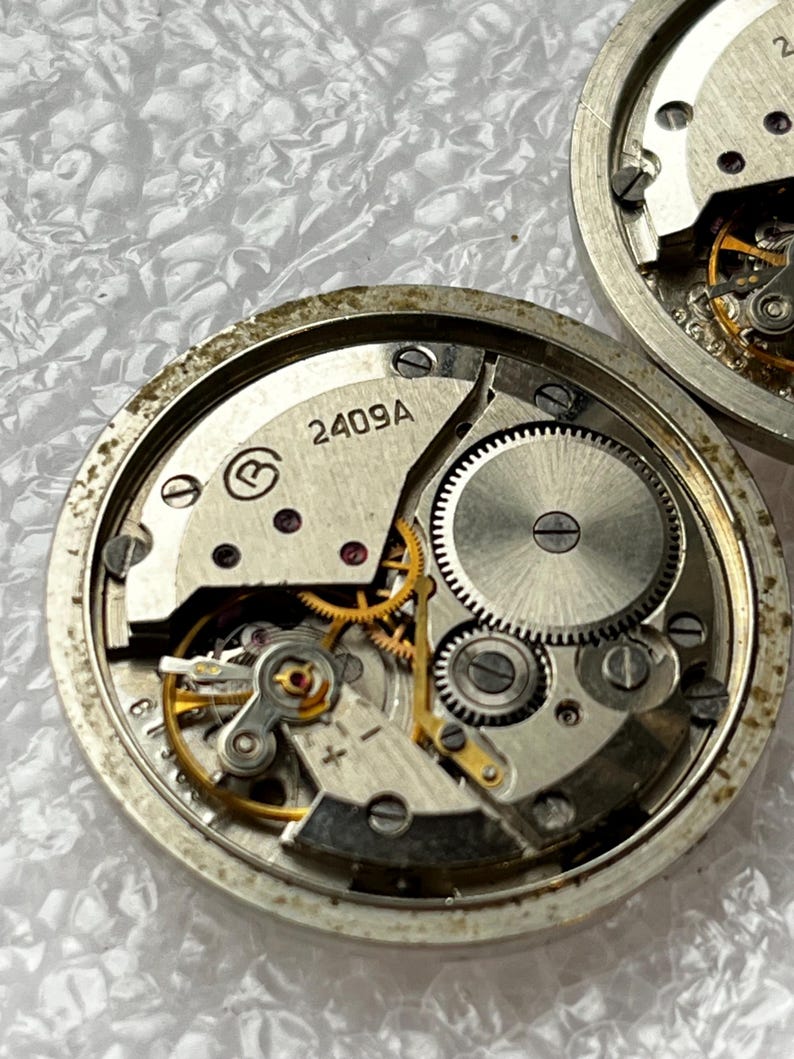 May include: Close-up of two disassembled watch movements. The watch mechanisms feature intricate gears, screws, and a balance wheel. The main plate is marked with "2409A". The components are silver and gold in color.