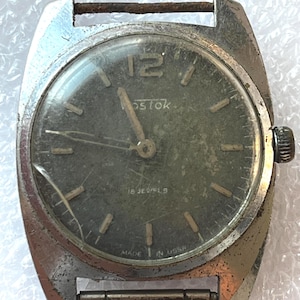 Può includere: Orologio da polso vintage con cassa usurata color argento e quadrante verde scuro. Il quadrante mostra il marchio "Vostok", il numero "12" e il testo "18 Jewels". L'orologio è contrassegnato "Made in USSR".
