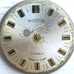 May include: Vintage Wostok watch face with a silver finish. It features gold and black rectangular hour markers and the text "Wostok", "17 Jewels", and "Made in USSR". The watch face is circular in shape.