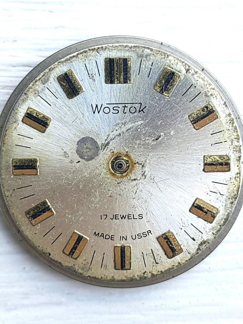 May include: Vintage Wostok watch face with a silver finish. It features gold and black rectangular hour markers and the text "Wostok", "17 Jewels", and "Made in USSR". The watch face is circular in shape.
