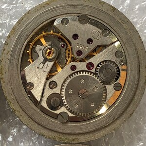 May include: Close-up view of a silver watch movement with intricate gears and a gold-coloured winding stem. The movement is labelled "2214".
