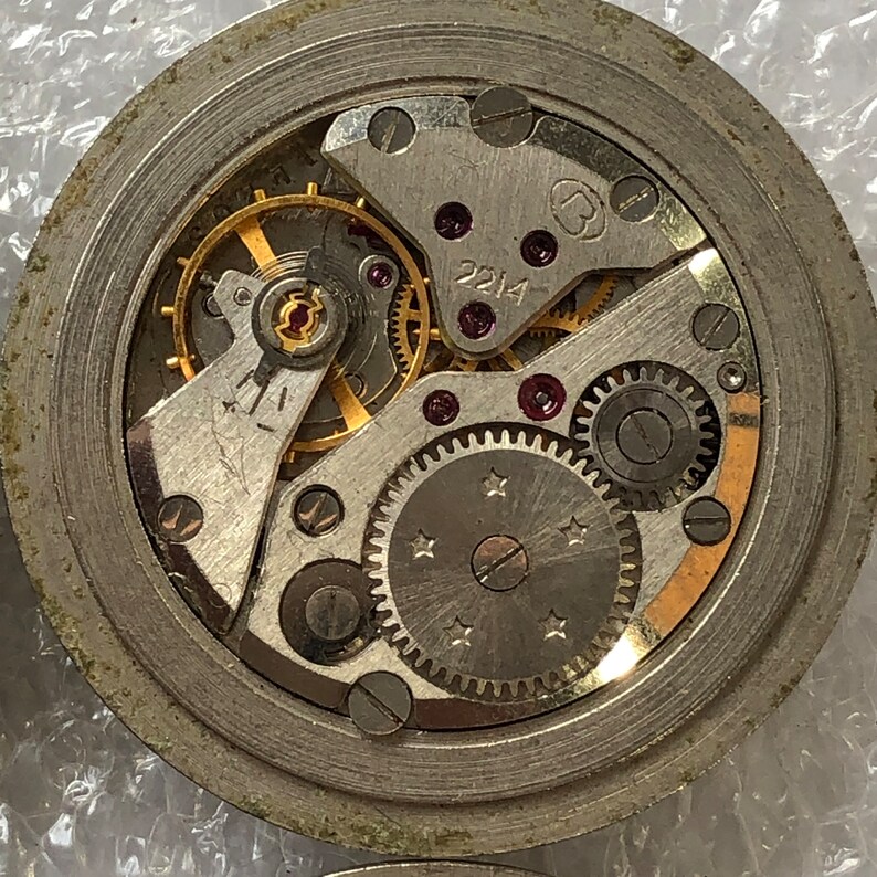 May include: Close-up view of a silver watch movement with intricate gears and a gold-coloured winding stem. The movement is labelled "2214".