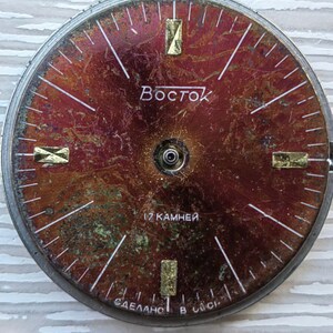 May include: A vintage watch face with a distressed, reddish-brown dial. The face features gold-colored rectangular hour markers and white lines indicating minutes. The word "Восток" is printed in Cyrillic script. The text "17 камней" and "СДЕЛАНО В СССР" are also visible.