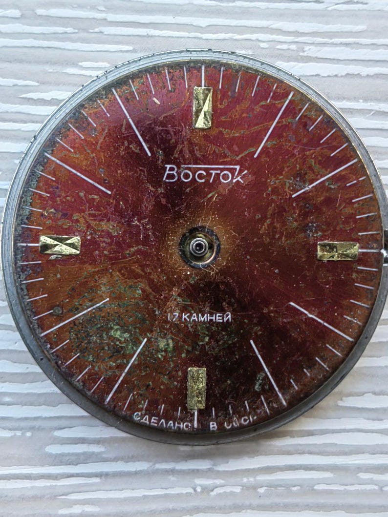 May include: A vintage watch face with a distressed, reddish-brown dial. The face features gold-colored rectangular hour markers and white lines indicating minutes. The word "Восток" is printed in Cyrillic script. The text "17 камней" and "СДЕЛАНО В СССР" are also visible.
