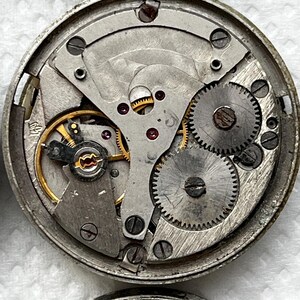 May include: Close-up of a disassembled watch mechanism. The intricate internal components, including gears, springs, and screws, are visible. The metal parts have a silver finish, with some gold-colored accents. The watch parts are arranged in a circular pattern.