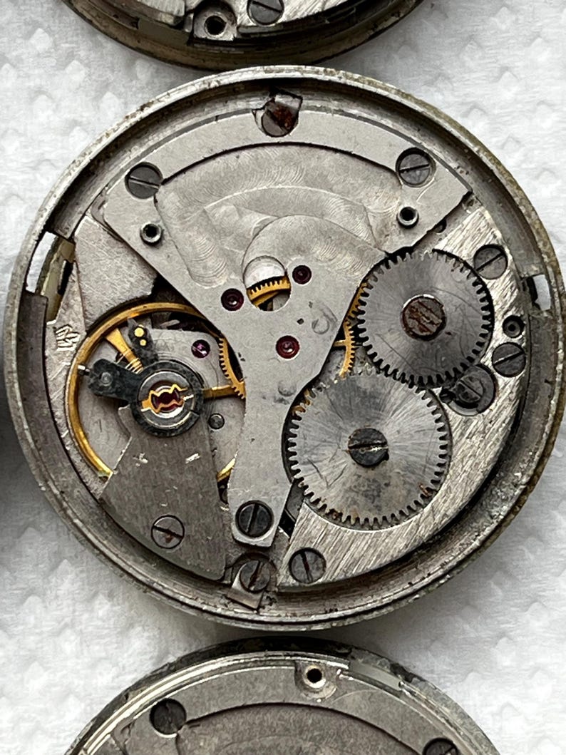 May include: Close-up of a disassembled watch mechanism. The intricate internal components, including gears, springs, and screws, are visible. The metal parts have a silver finish, with some gold-colored accents. The watch parts are arranged in a circular pattern.