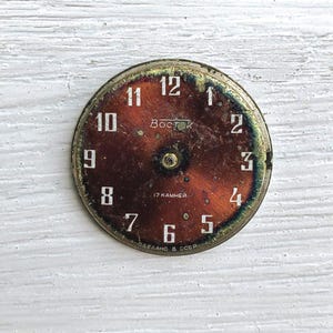 May include: A vintage watch face with a rusty brown dial and white numerals. The face has the text "Восток" and "17 KAMHEЙ". The watch face is round with a gold-colored metal rim.