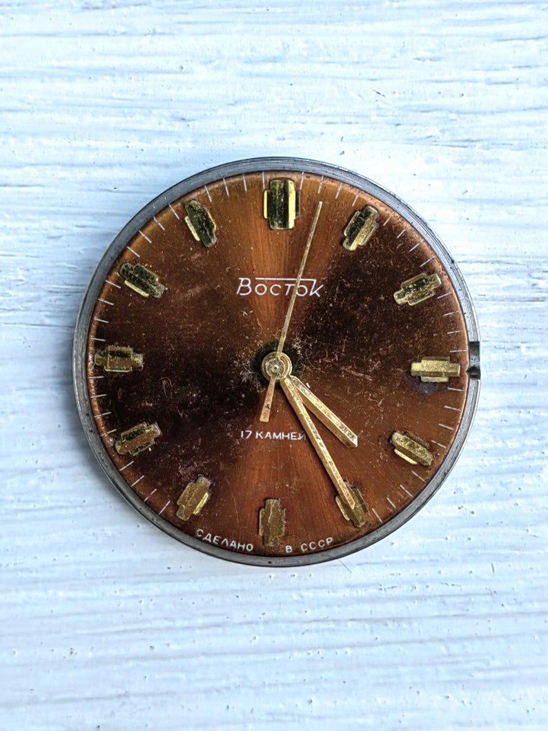 May include: A vintage watch face with a copper-colored dial and gold-toned hour markers. The watch face has the word "Восток" in white lettering and the text "17 камней" and "Сделано в СССР".