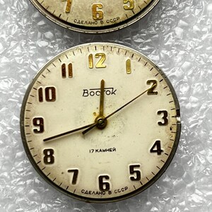 May include: Two vintage watch faces with cream-colored dials and gold-toned hands and numerals. The top face shows the numbers 4, 5, 6, 7, and 8. The bottom face has the word "Bocrok" and the numbers 1 through 12.