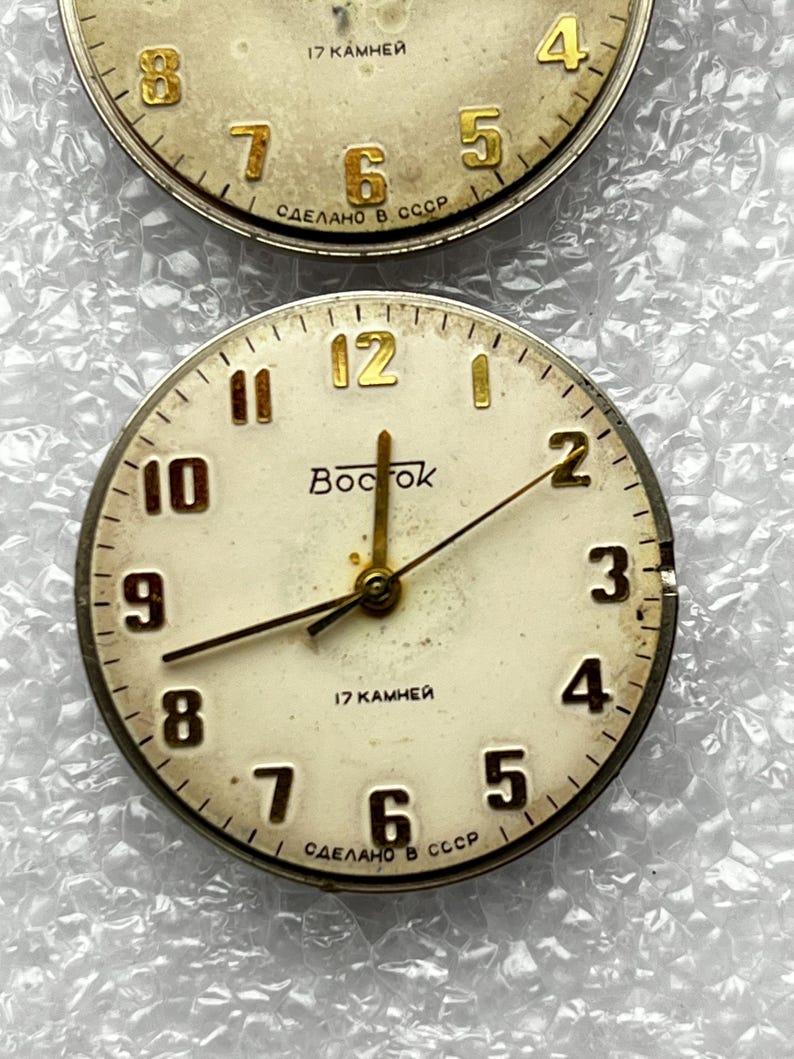 May include: Two vintage watch faces with cream-colored dials and gold-toned hands and numerals. The top face shows the numbers 4, 5, 6, 7, and 8. The bottom face has the word "Bocrok" and the numbers 1 through 12.