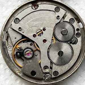 May include: Close-up of a vintage watch movement, showcasing intricate gears, screws, and jewels. The silver-toned metal components are arranged within a circular frame. The image highlights the mechanical complexity of the watch.
