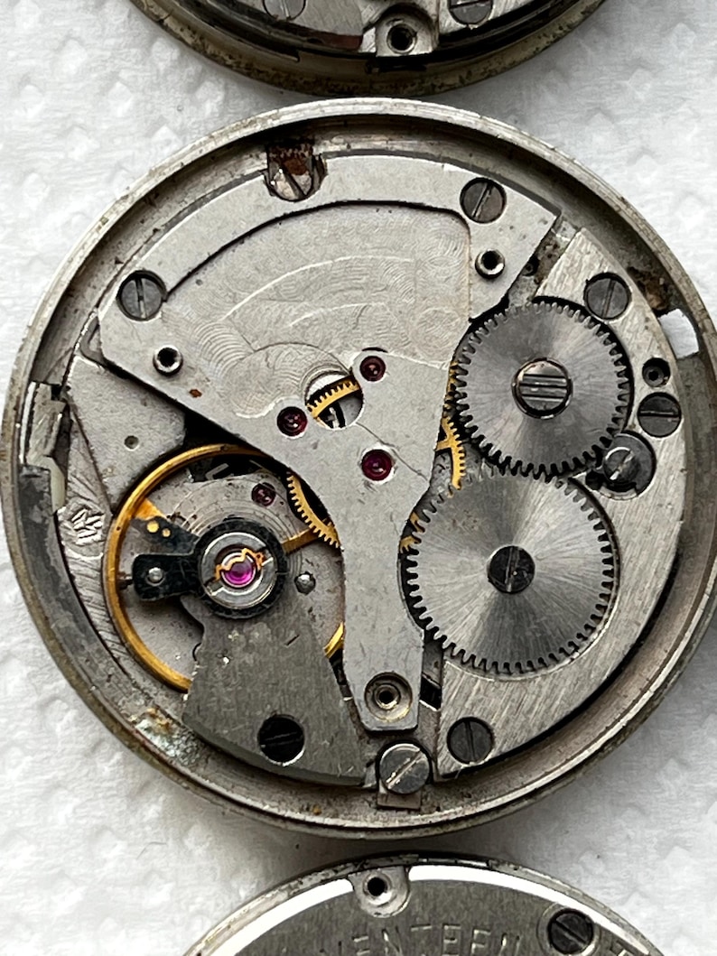 May include: Close-up of a vintage watch movement, showcasing intricate gears, screws, and jewels. The silver-toned metal components are arranged within a circular frame. The image highlights the mechanical complexity of the watch.