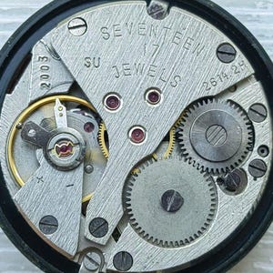 May include: Close-up of a watch movement with visible gears, screws, and the text "SEVENTEEN 17 JEWELS". The metal components are silver and gold, set within a black circular frame. The watch mechanism is intricate and detailed.