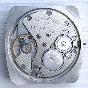May include: Close-up of a vintage watch movement with visible gears, screws, and the text "SEVENTEEN 17 JEWELS". The metal components are silver-toned, with red jewels and gold accents. The watch movement is square-shaped.