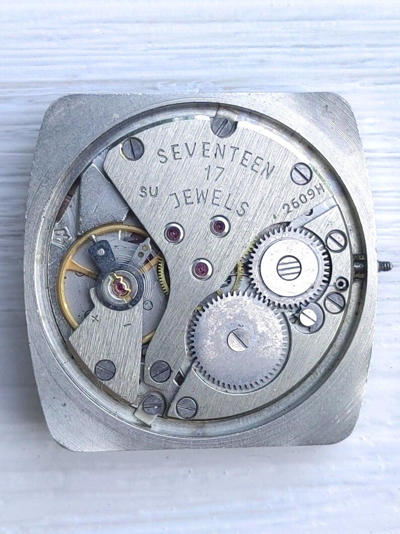 May include: Close-up of a vintage watch movement with visible gears, screws, and the text "SEVENTEEN 17 JEWELS". The metal components are silver-toned, with red jewels and gold accents. The watch movement is square-shaped.