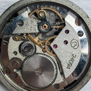 May include: Close-up of a vintage watch movement, showcasing intricate gears, springs, and other mechanical components. The watch's internal mechanism is visible, with a silver and gold color scheme. The watch face is not present, revealing the inner workings.