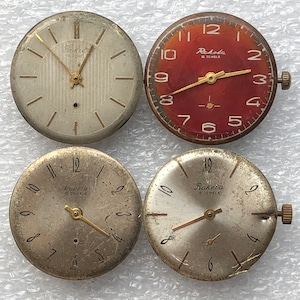May include: Four vintage watch faces, two with silver colored faces and black numbers, one with a red face and black numbers, and one with a silver face and black numbers. All faces have gold colored hands and the text "Rakela" and "15 Jewels" or "16 Jewels".