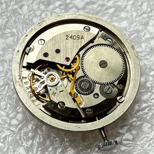 May include: Close-up of a watch movement with visible gears, springs, and other internal components. The metal frame is circular, with the text "2409A" visible. The watch mechanism is silver and gold in color.