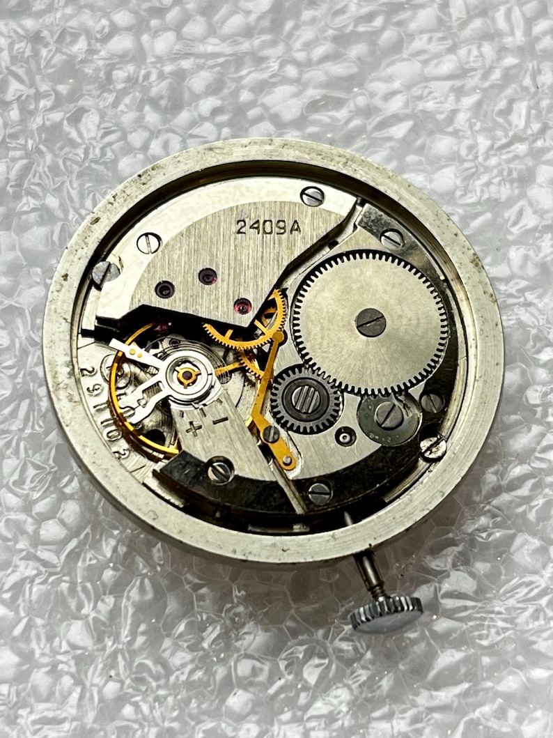 May include: Close-up of a watch movement with visible gears, springs, and other internal components. The metal frame is circular, with the text "2409A" visible. The watch mechanism is silver and gold in color.