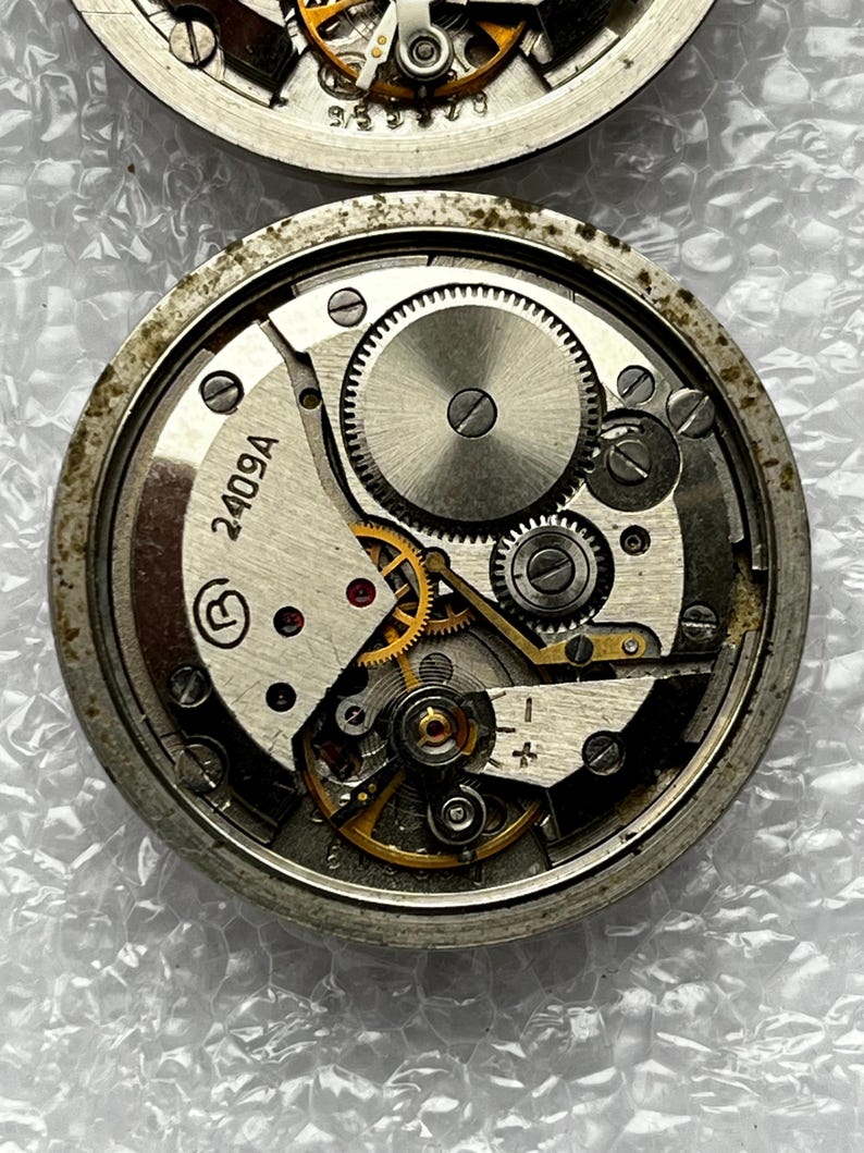 May include: Close-up of two watch movements, showcasing intricate gears and mechanisms. The lower movement features the number "2409A" and a circular symbol. The gears are silver and gold, set within a circular metal frame.