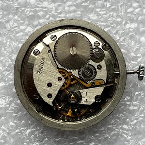 May include: Close-up of a disassembled watch movement. The silver-colored mechanism features numerous gears, springs, and other components. The watch movement has a winding stem and the number "2409A" is visible.